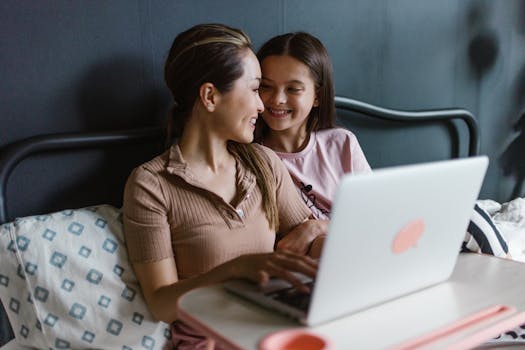 A mother and daughter enjoy quality time together on a bed while using a laptop. Perfect work-from-home moment.
