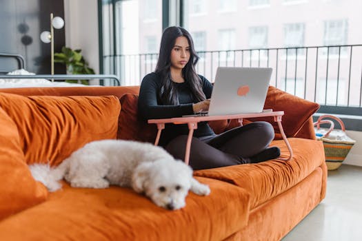Woman sitting on a sofa with a laptop, accompanied by a dog indoors.