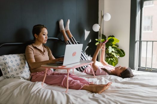 A mother works on a laptop while her daughter relaxes with a phone on a cozy bed.
