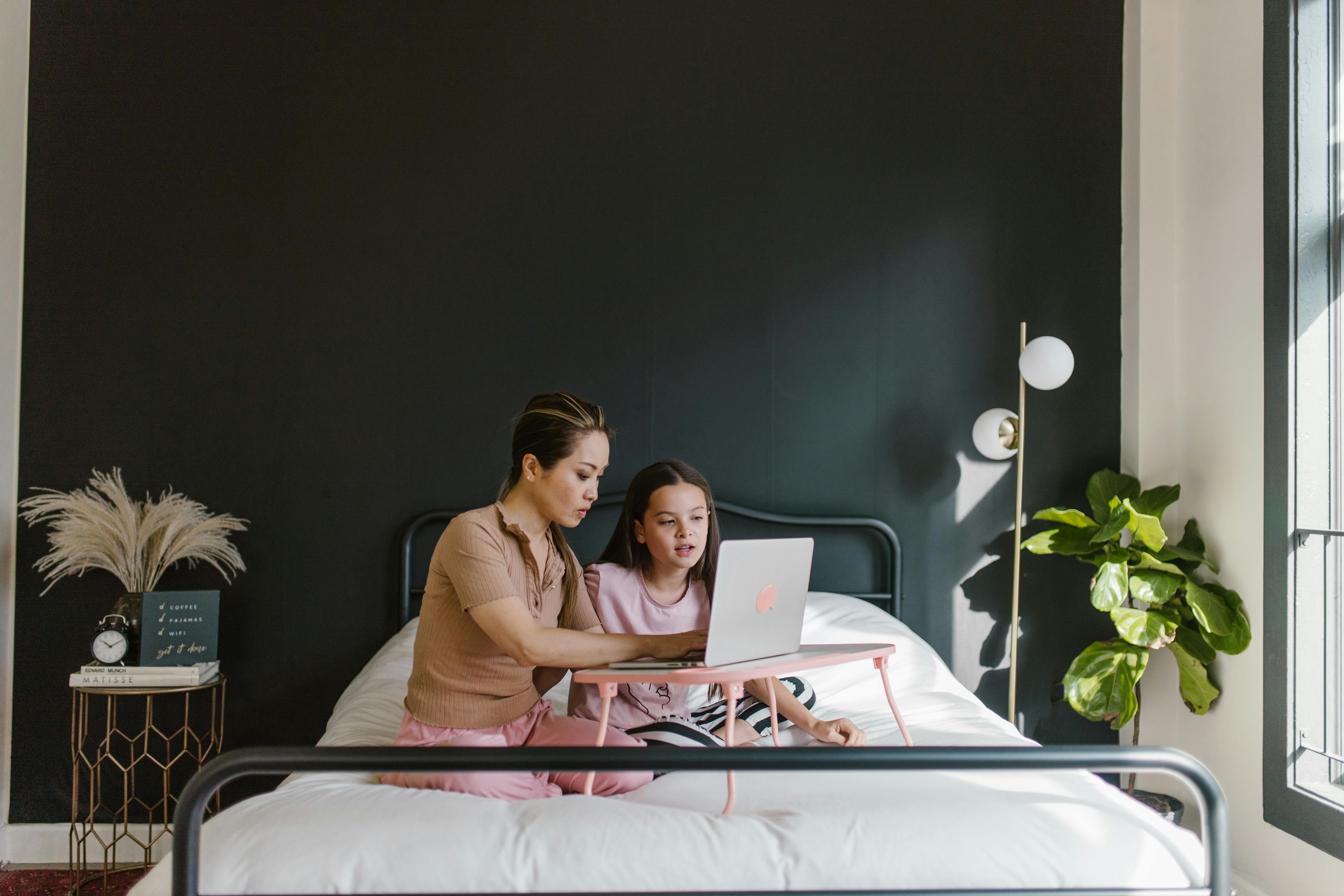 A mother and daughter sit together on a bed using a laptop during the day, promoting togetherness.
