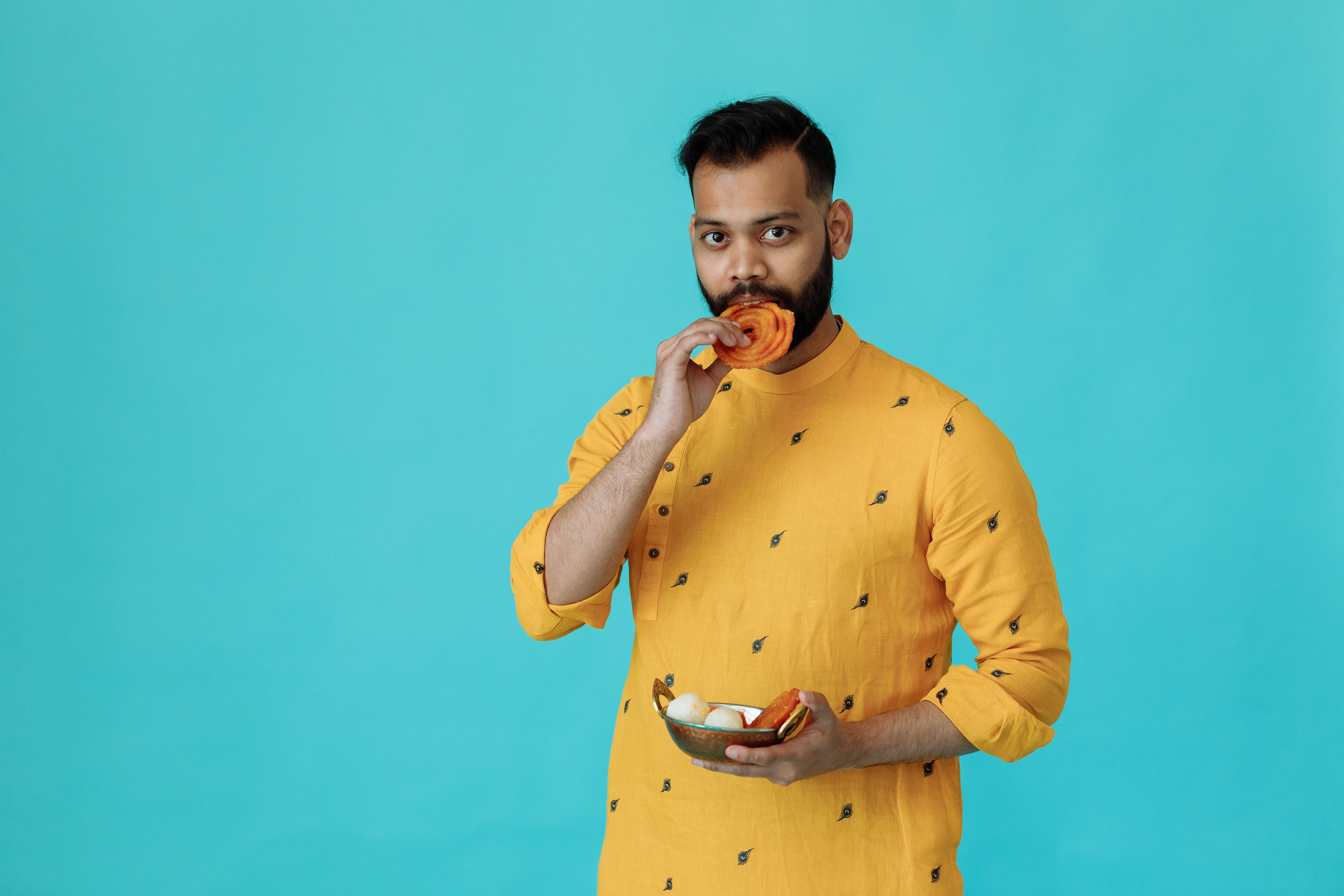 Indian man in traditional attire eating sweets in a studio setting.
