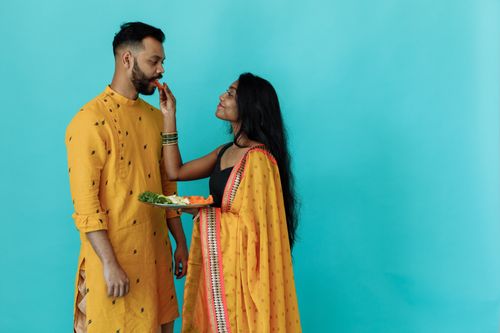 Free An Indian couple in traditional yellow attire shares a playful moment with food against a blue background. Stock Photo