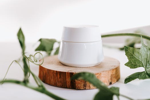 A simple white skincare container atop a wood slice surrounded by fresh green leaves.