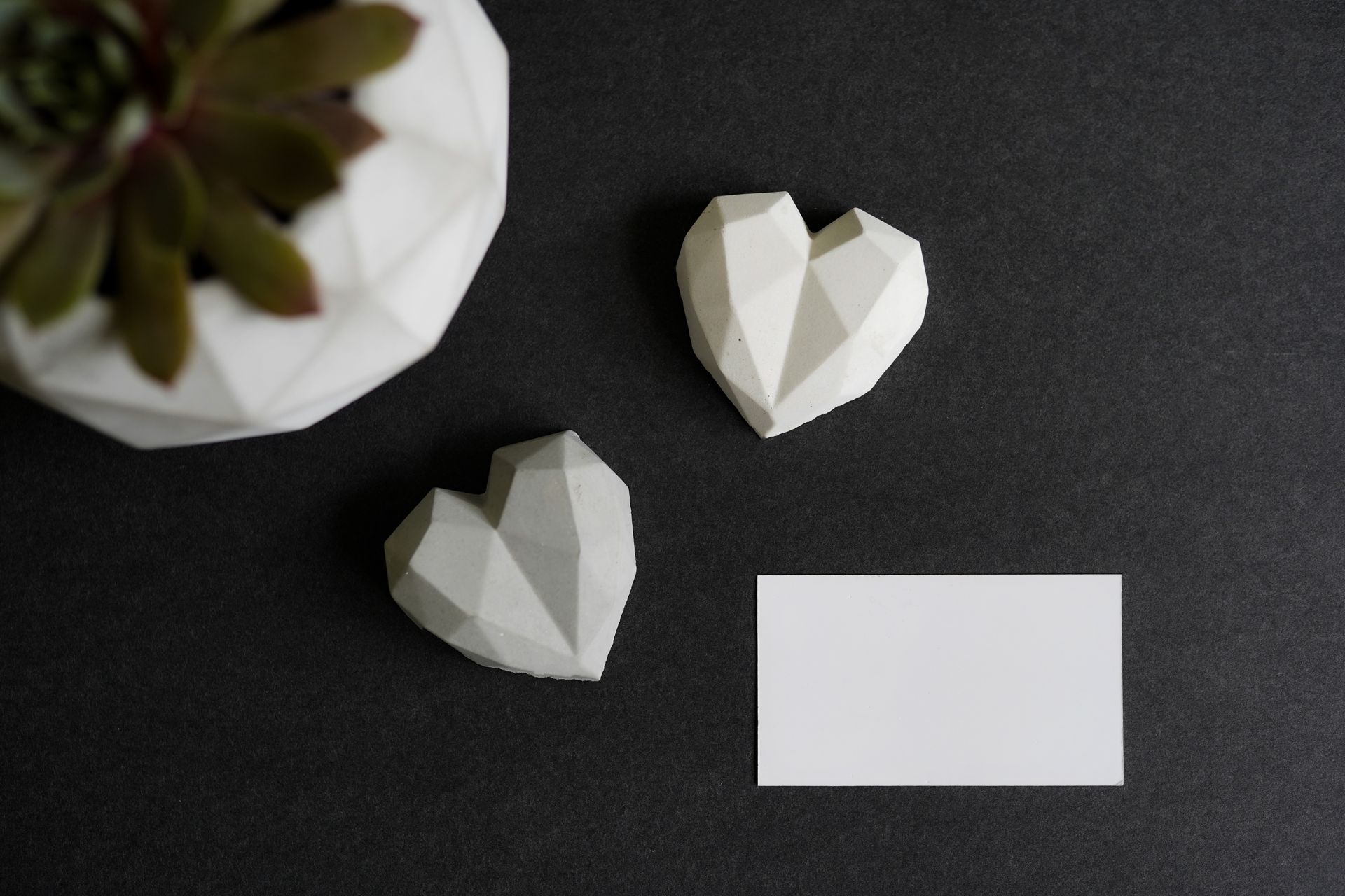 A minimalist flat lay featuring geometric hearts, a succulent in a white planter, and a blank business card on a dark background.