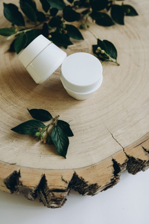Free White cosmetic containers placed on a wooden slab with green leaves, creating a natural skincare theme. Stock Photo