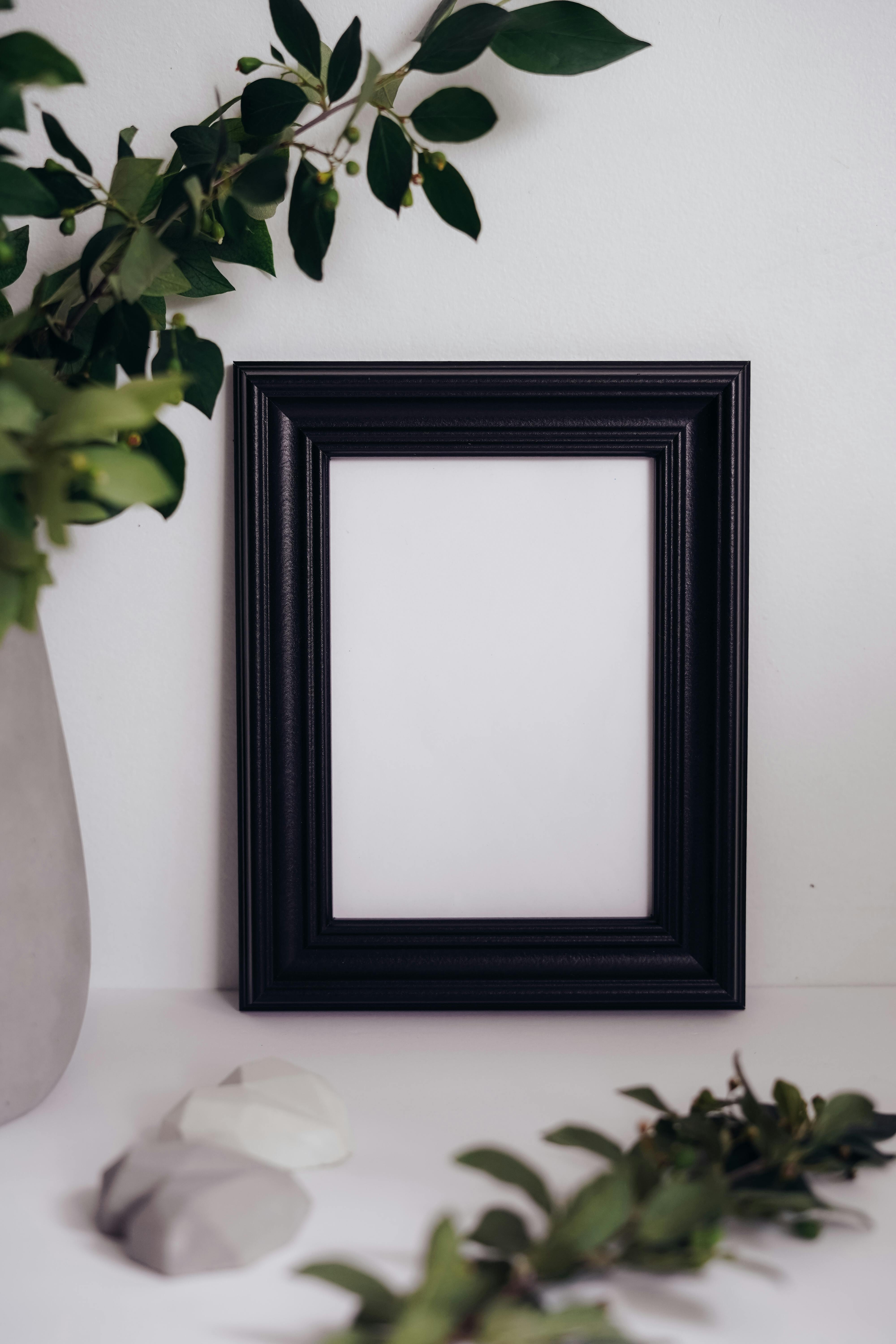 Stylish black frame with green leaves and elegant stones against a white wall, perfect for mockup designs.