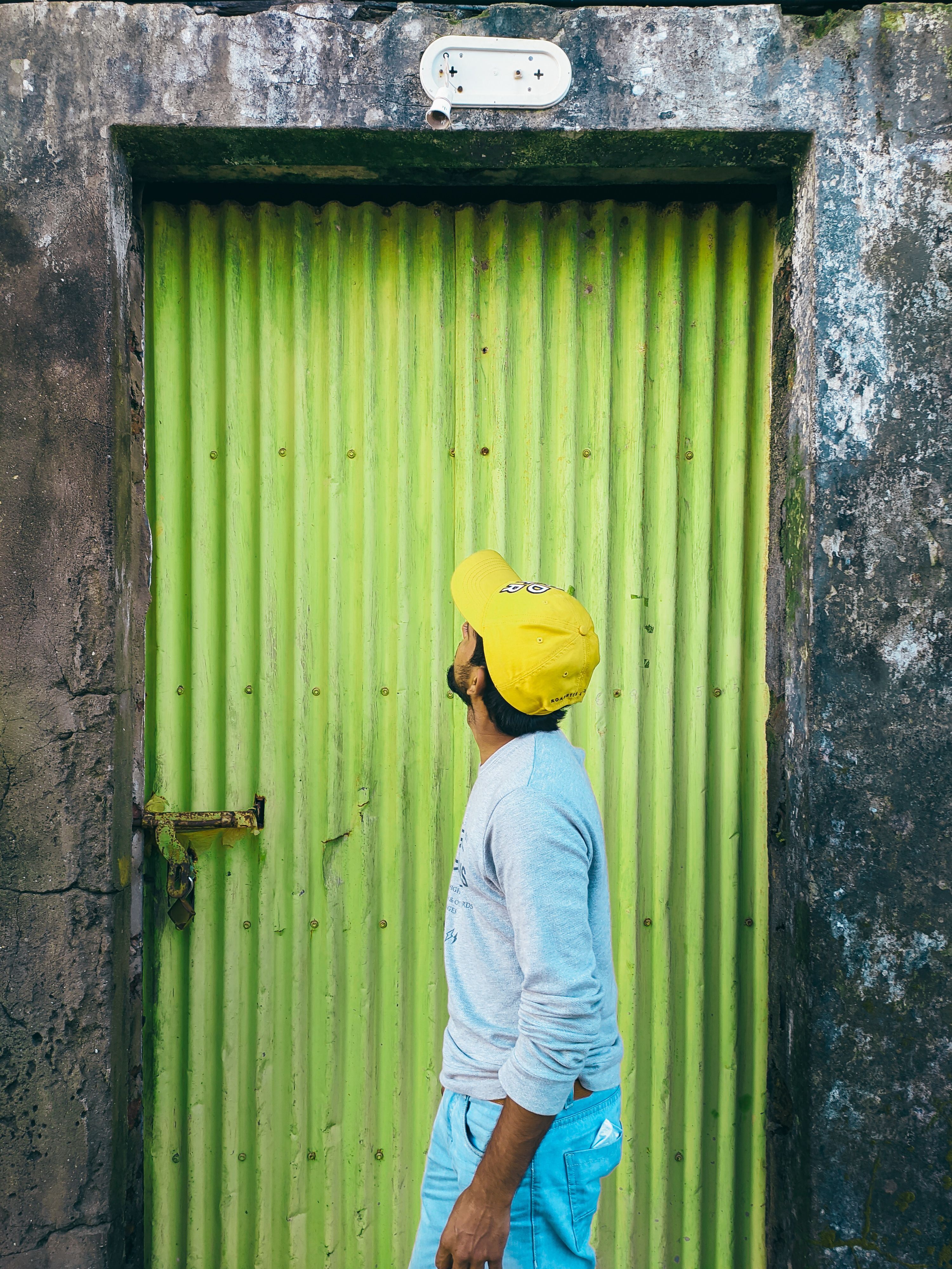 A person wearing a yellow cap looks up at a textured green door, standing in a vertical frame.