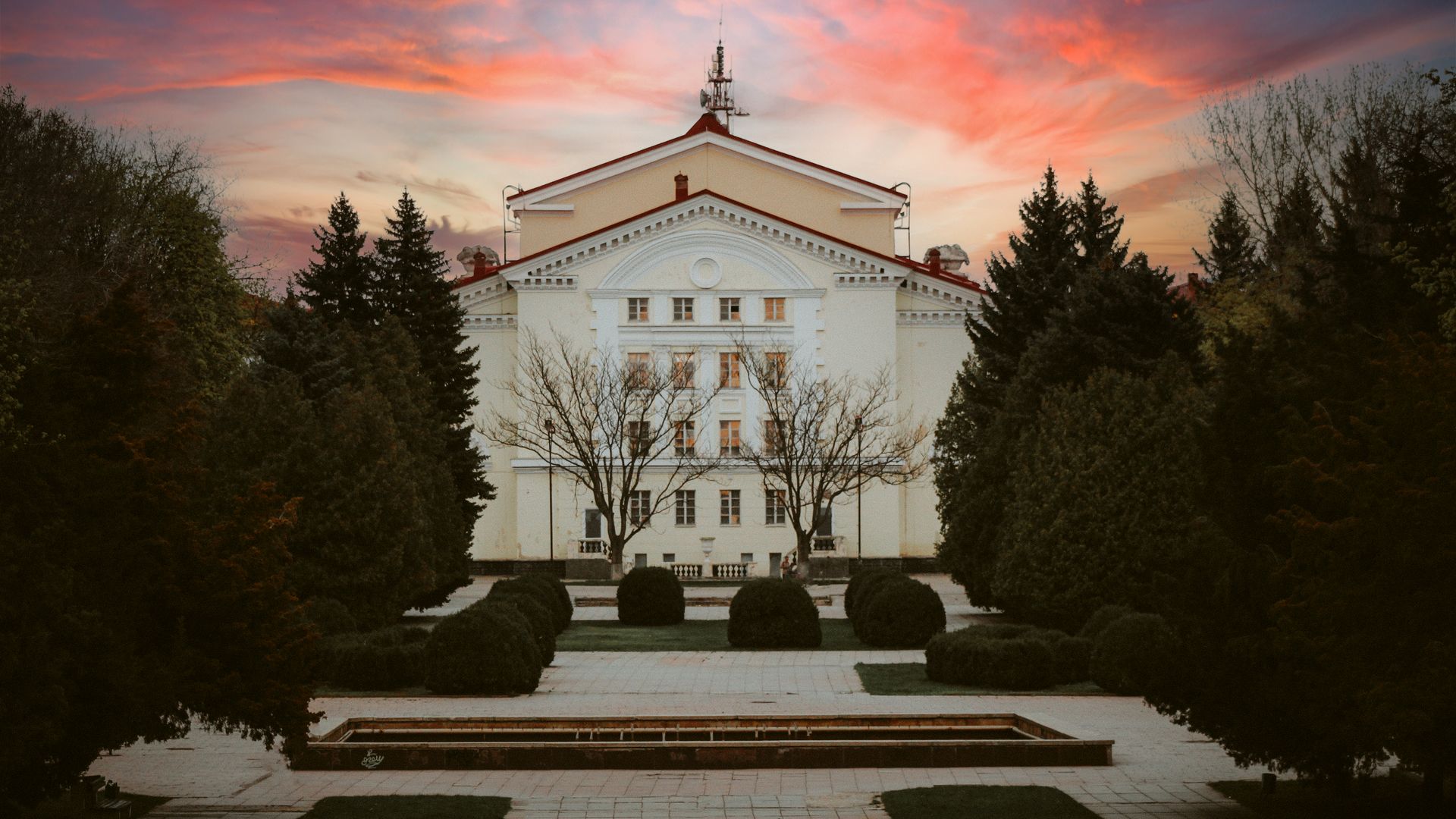 A grand mansion surrounded by trees and gardens at sunset with vibrant skies.