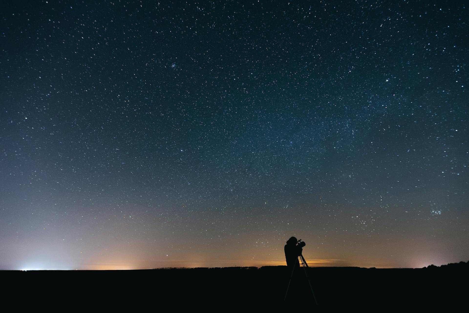 A person using a telescope under a vast, star-filled night sky with a silhouette effect.