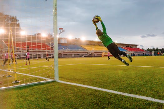 Goalkeeper making a diving save on a soccer field during a match at sunset.