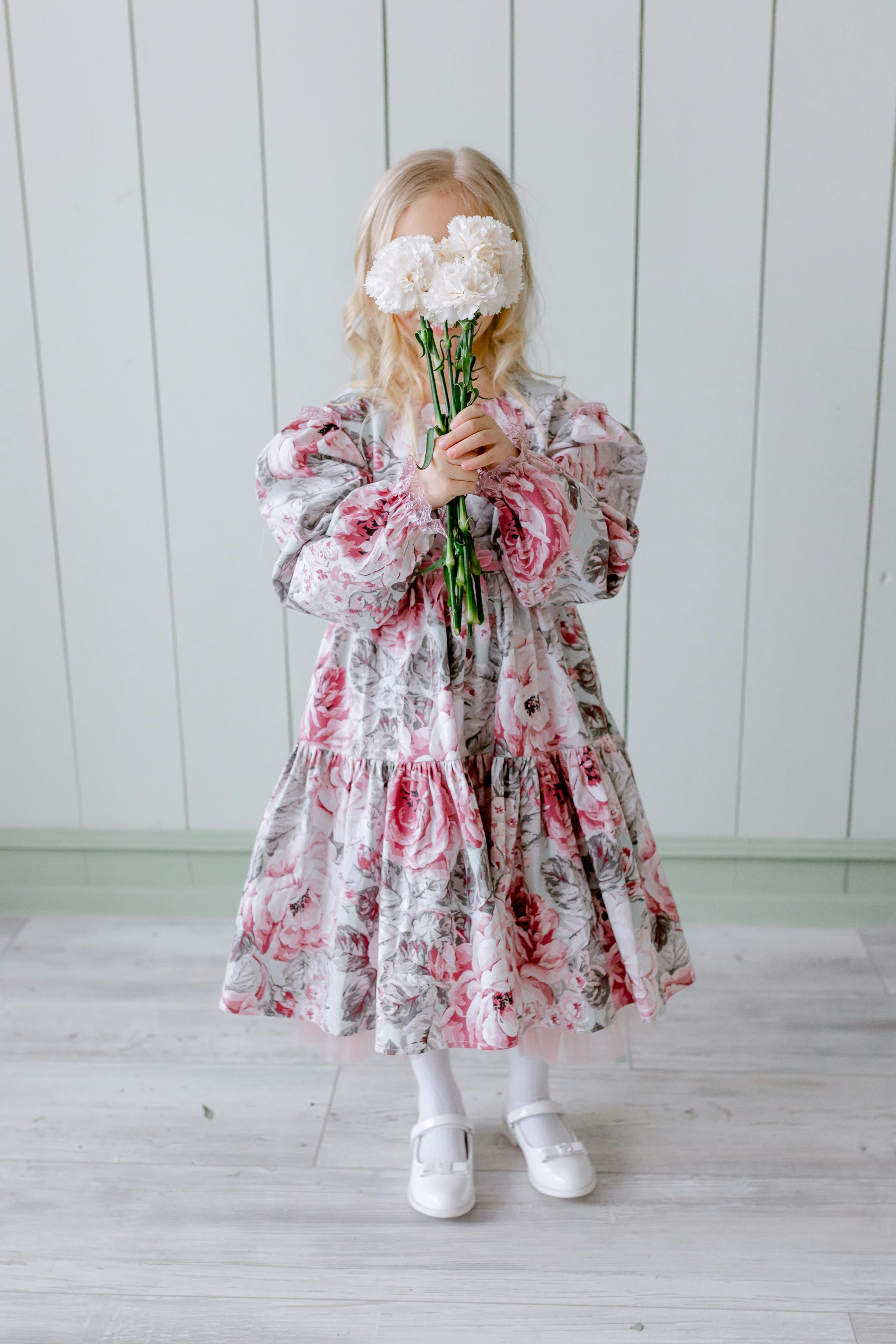 A little girl in a floral dress holds white flowers, standing indoors on a light wooden floor.