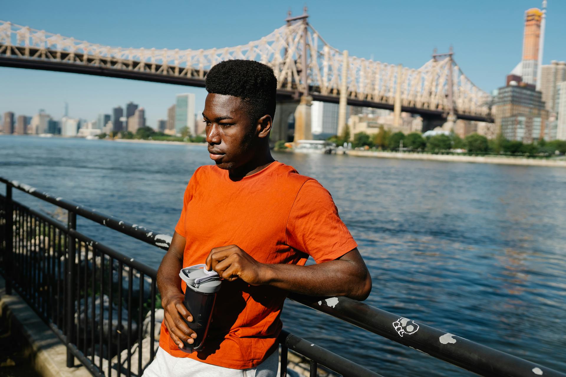 A young man in an orange shirt leans on a railing by the East River, holding a tumbler with a bridge in the background.