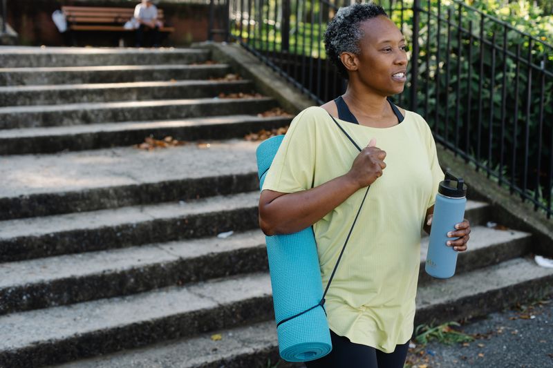 A Black woman with closely cropped gray hair, in a yellow t-shirt, walks down concrete park steps carrying a rolled teal yoga mat on her shoulder and a light blue water bottle.