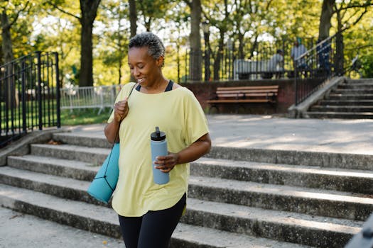African American woman in activewear walking outdoors with a yoga mat and water bottle.