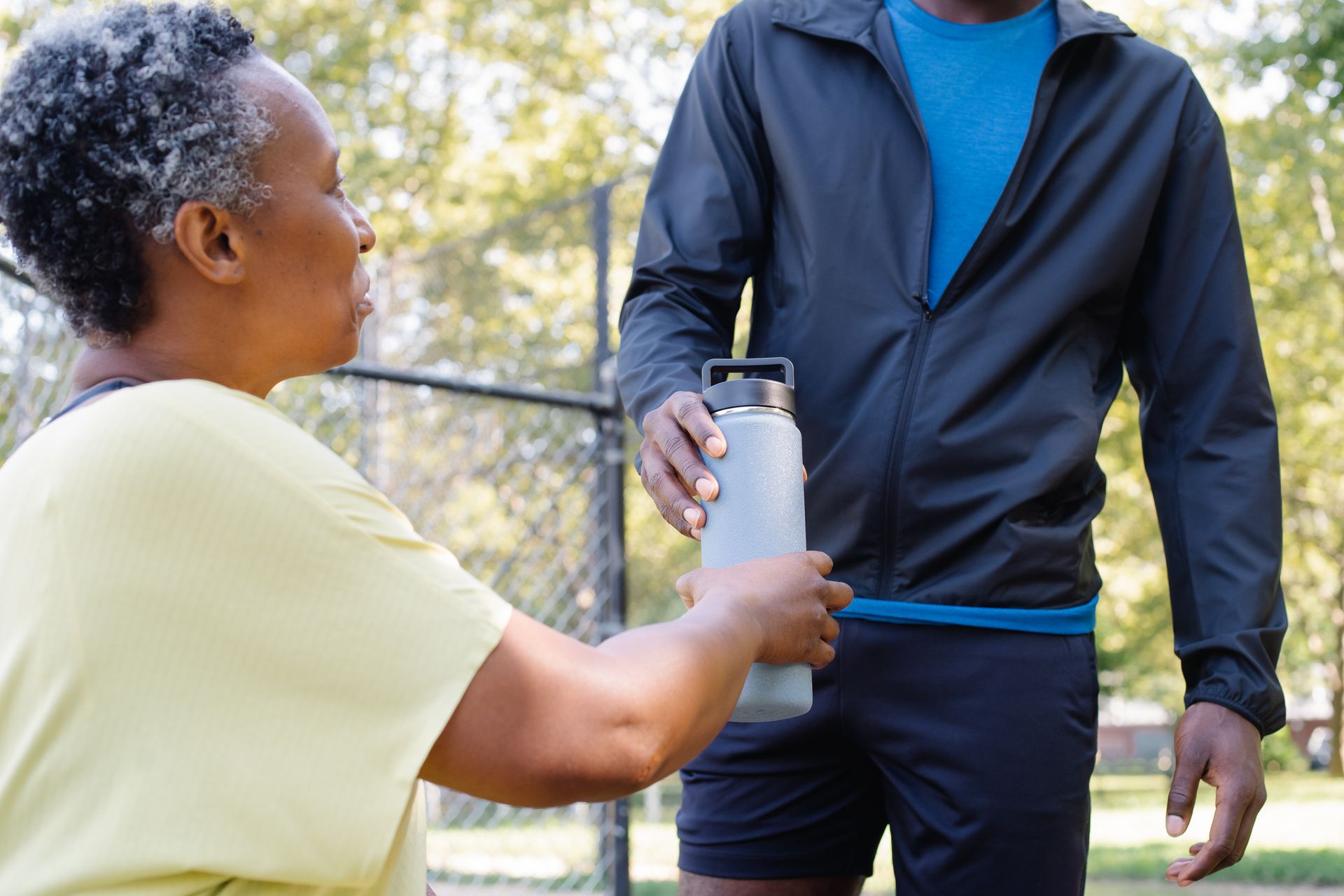 An adult woman and man share a water bottle in a sunny park, emphasizing bonding and fitness.