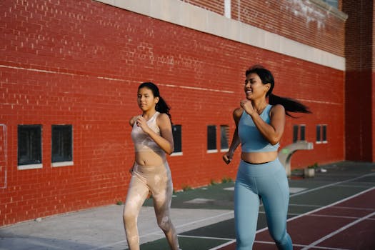 Two women in sportswear jogging outdoors on a sunny day beside a red brick wall.