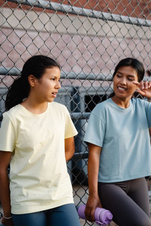 Free Two teenagers in casual T-shirts stand by a chain link fence, engaged in conversation outdoors. Stock Photo