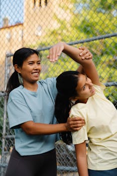 Woman guiding another in a stretching routine outdoors by a fence.