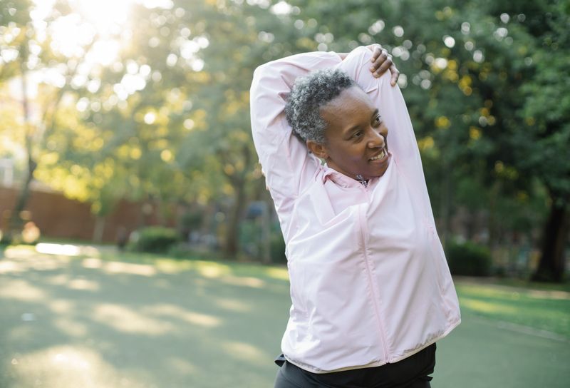 An older Black woman in a pink jacket stretches her arms during an outdoor workout in a green park, the kind of weight-bearing activity that protects aging bones.
