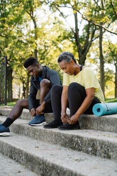 Two adults preparing for outdoor exercise, tying shoes on concrete stairs with a yoga mat nearby.