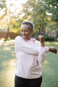 Happy woman in pink jacket stretching arms in sunny park setting.