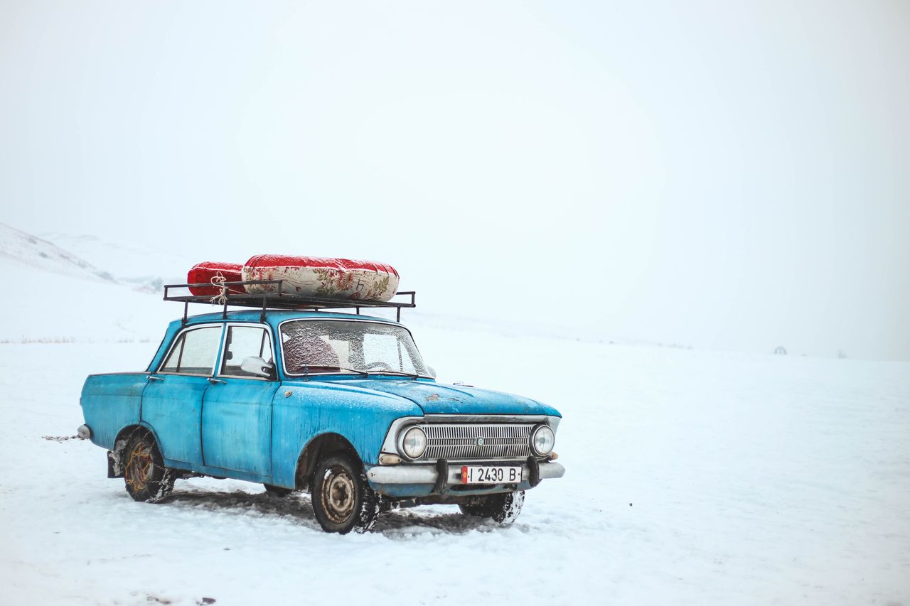 a vintage blue car with luggage on top amidst a snowy landscape in kyr