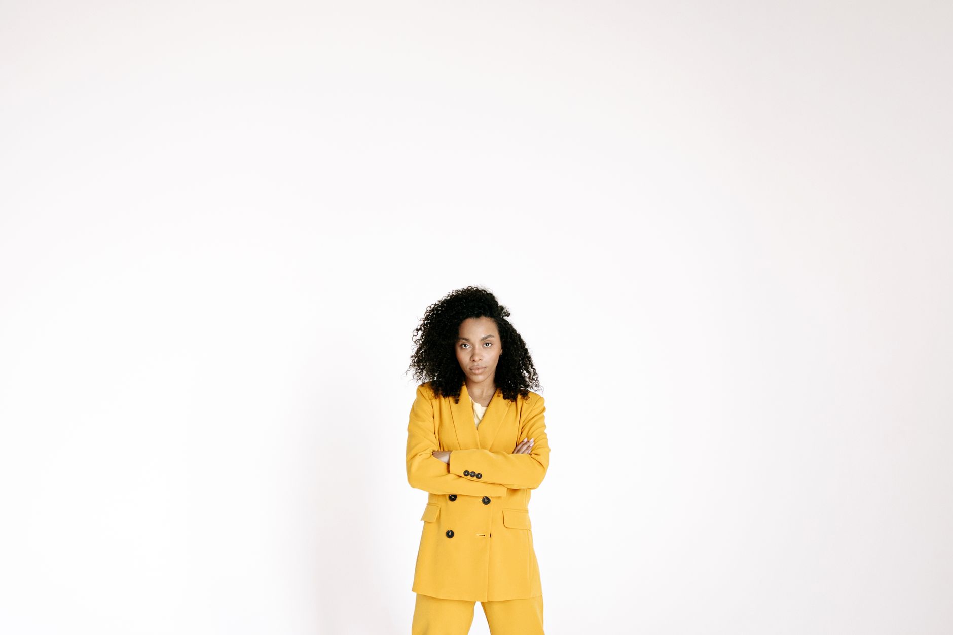 A confident woman with curly hair in a yellow suit, arms crossed, standing in a studio