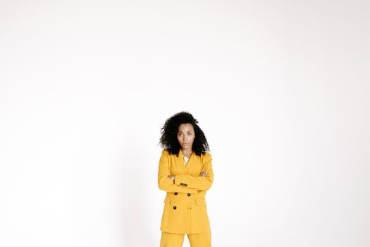 Portrait of a confident woman in a yellow suit with curly hair, arms crossed, posing in a studio setup.