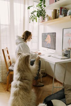 A woman works at a computer desk indoors with her dog standing beside her in a cozy home setting.