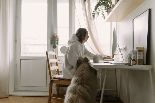 A woman in a white hoodie works on a computer at home with her dog beside her.