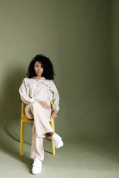 Confident young woman with curly hair sitting on a yellow chair against a green backdrop.
