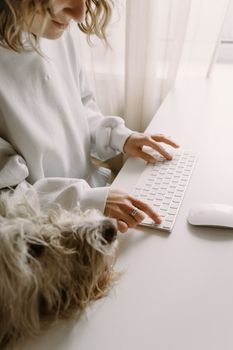 A woman in a white sweater typing on a keyboard with her furry dog companion beside her.