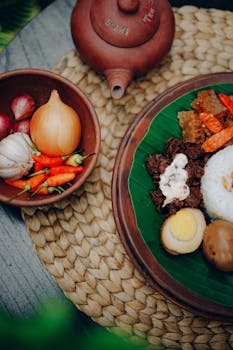 Top-down view of authentic Indonesian dishes with fresh ingredients and traditional teapot on woven mat.