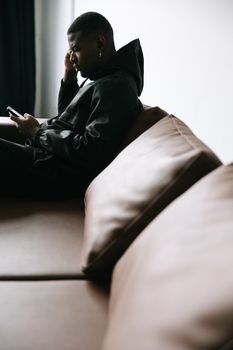 Side view of a man sitting on a couch indoors, using a smartphone. Modern lifestyle concept.
