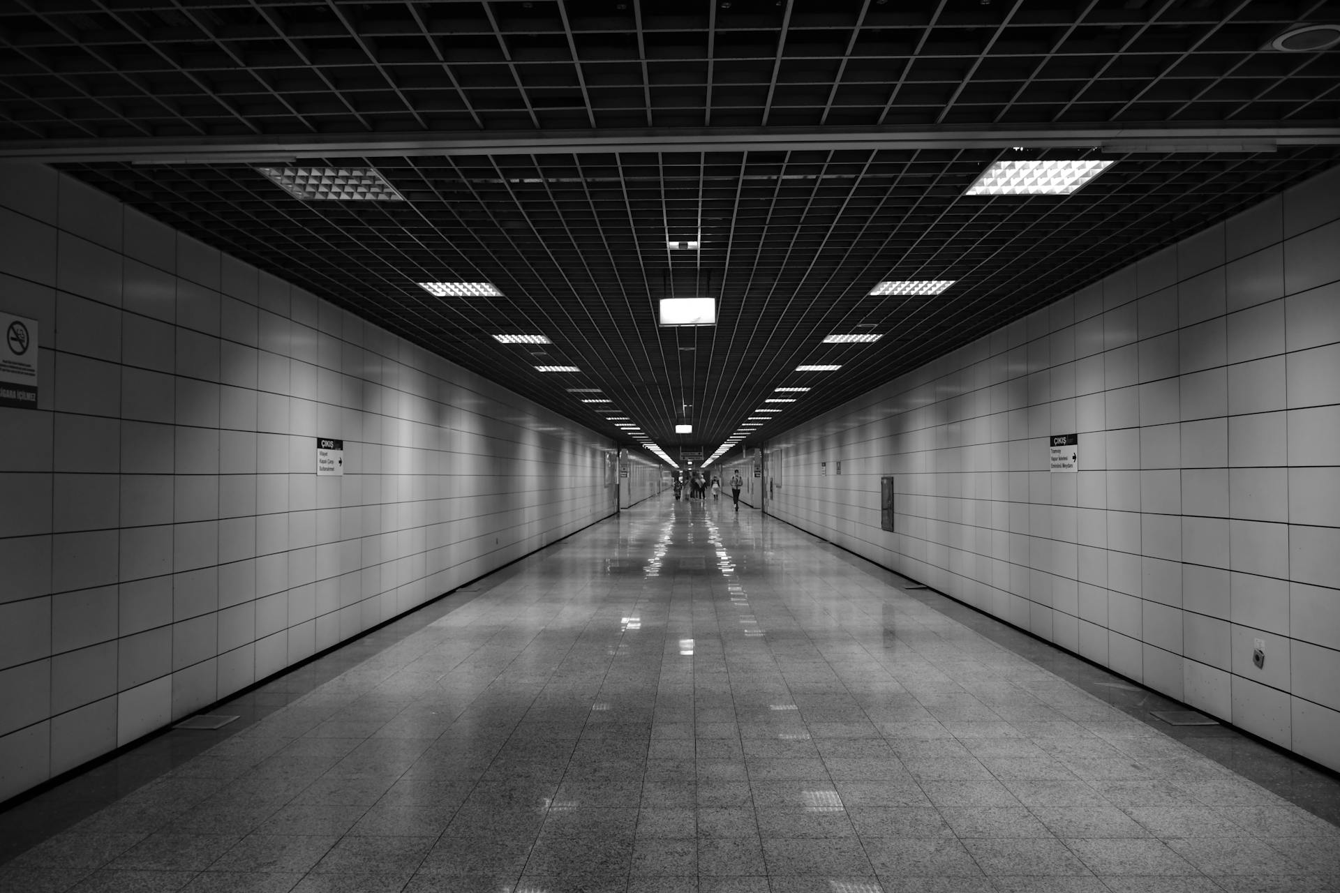 Black and white perspective view of a long, empty underground hallway with tiled walls and ceiling lights.