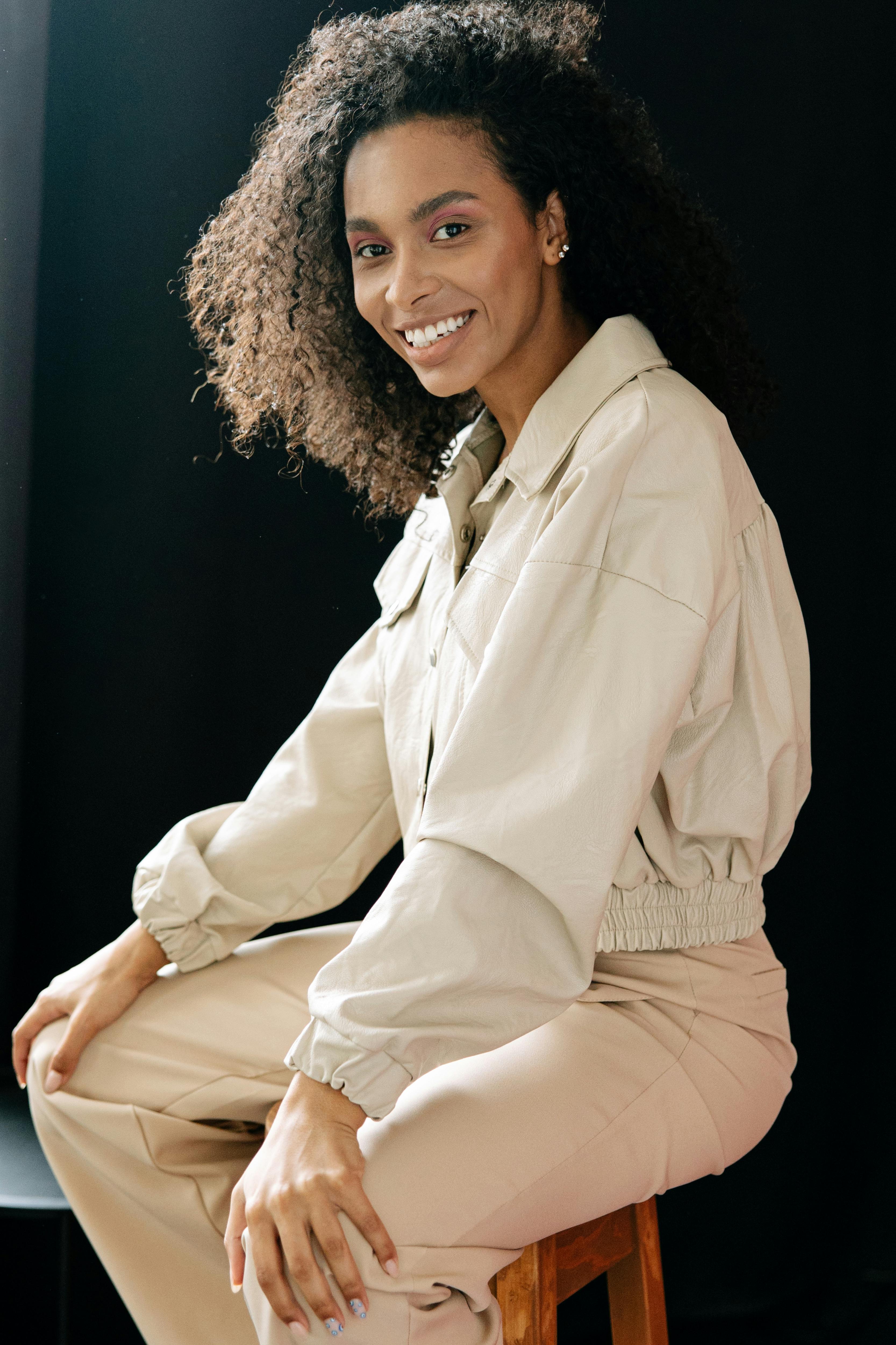 Fashionable woman with curly hair sitting and smiling in a beige outfit against a dark background.