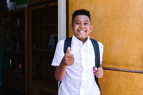 Free Smiling student with backpack giving thumbs up, standing at school entrance. Stock Photo