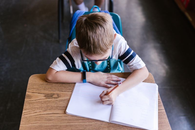 A young boy wearing glasses writing in a notebook at a school desk.