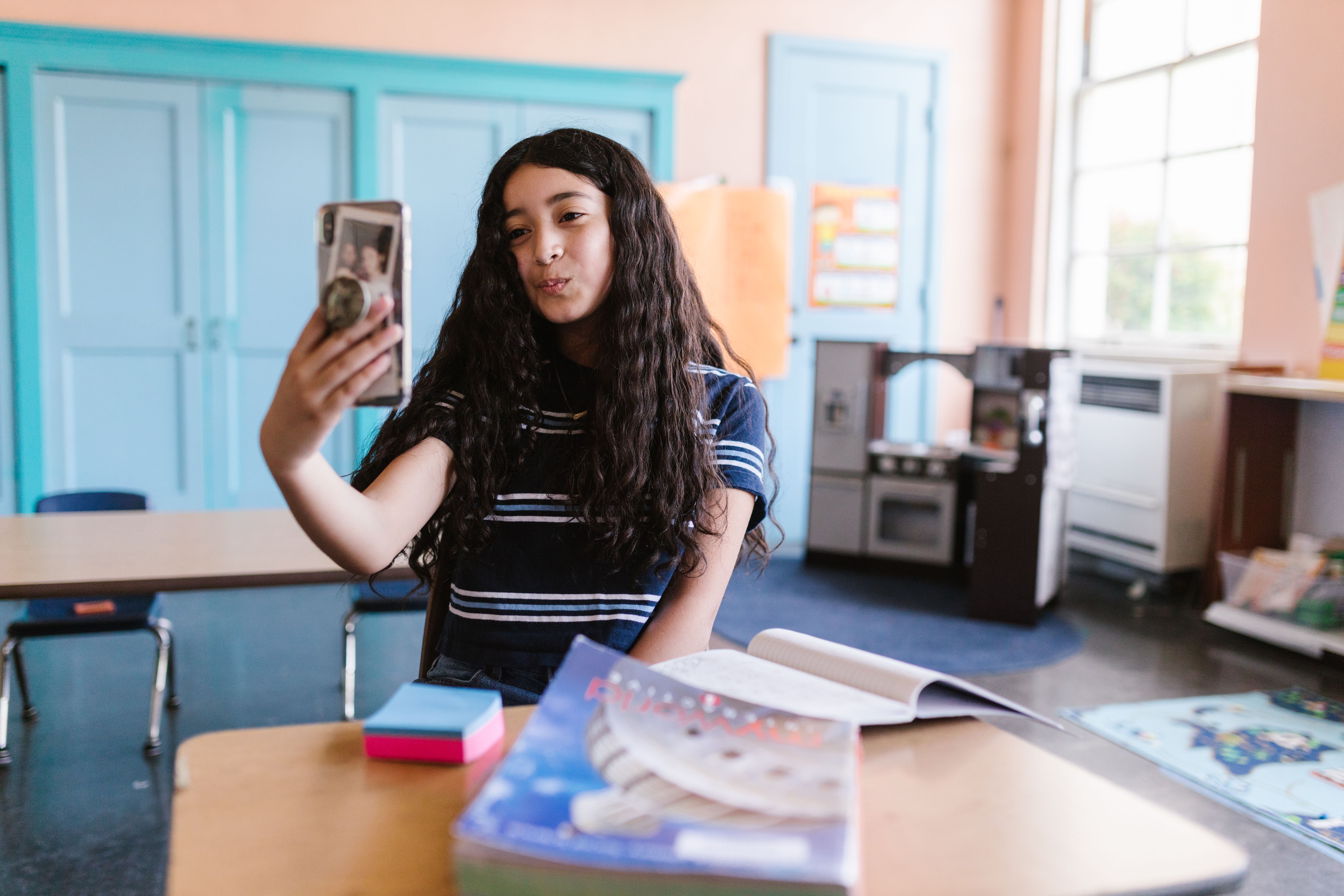 A teenage girl captures a selfie in a colorful classroom, surrounded by books and supplies.