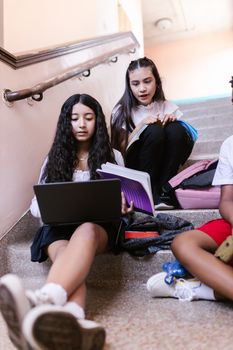 A group of teenagers studying together on a school staircase with laptops and books.