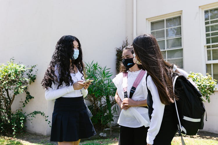 Schoolgirls Wearing Backpacks And Face Masks Talking In Front Of School Building