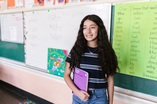 Young girl in a classroom holding a notebook, smiling at the camera with a backdrop of educational materials.