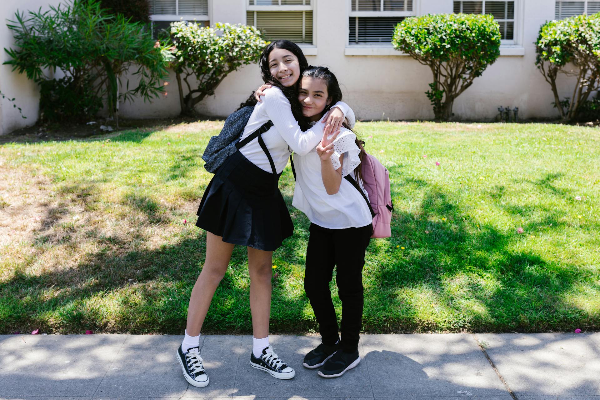 Two friends hugging outside a school building on a sunny day, both wearing backpacks.