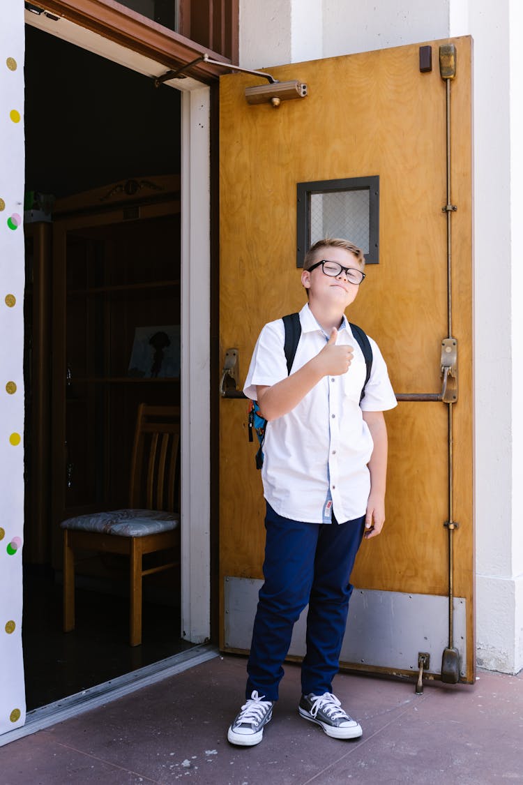 Boy In Blue And White School Uniform Standing Beside A Wooden Door With Thumbs Up