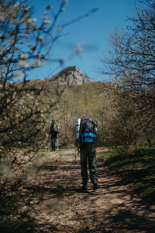 Two People Walking on Dirt Road Near Mountain Under Blue Sky