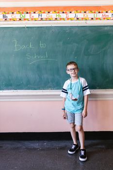 Smiling young boy standing in classroom with 'Back to School' written on chalkboard.