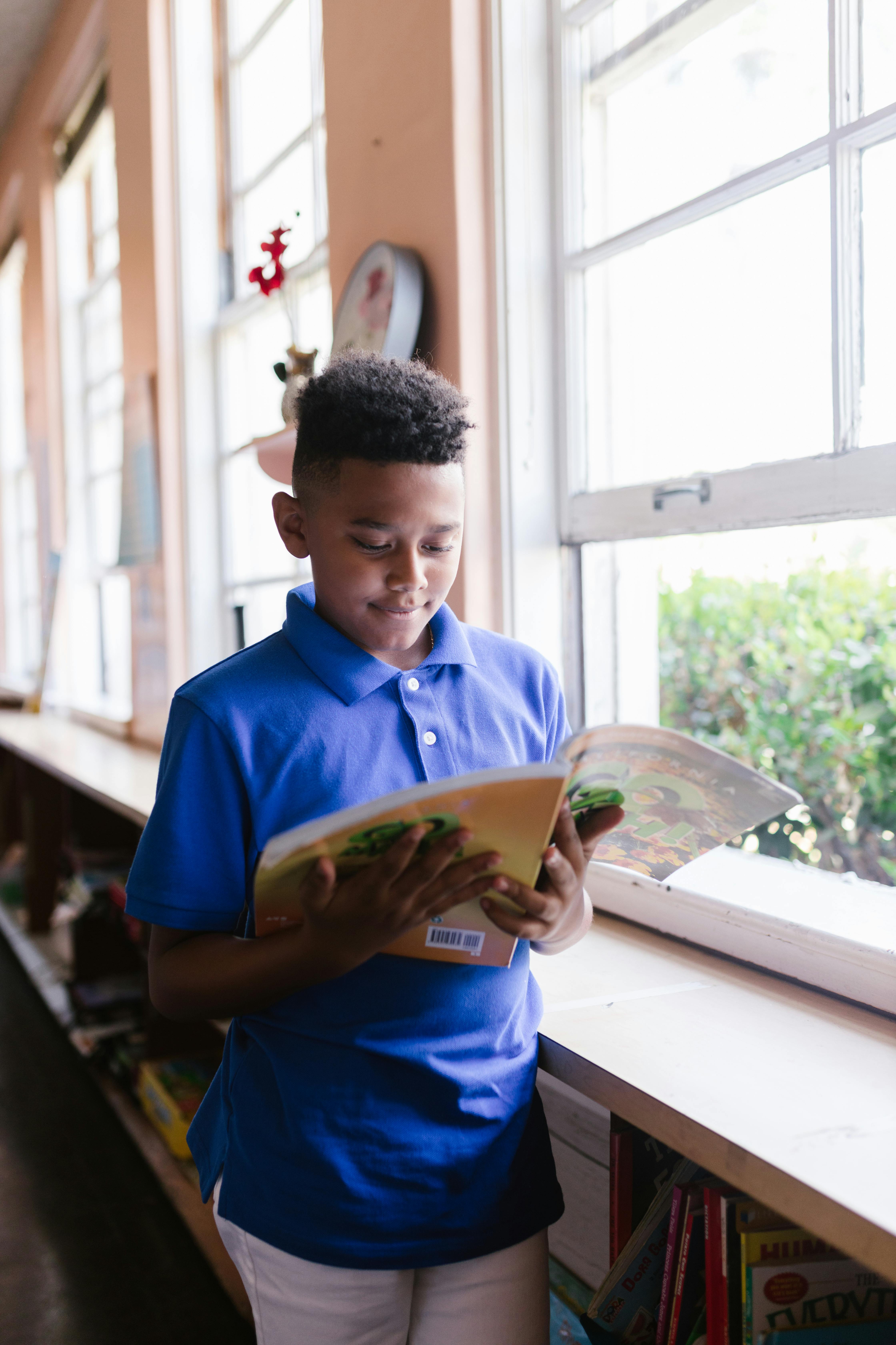 Free A boy in a blue polo reads a book by a sunlit window, enjoying a quiet moment indoors. Stock Photo