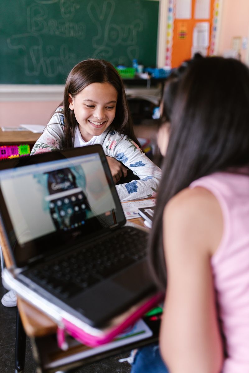 Two girls in a classroom working together on a digital school project with a laptop.