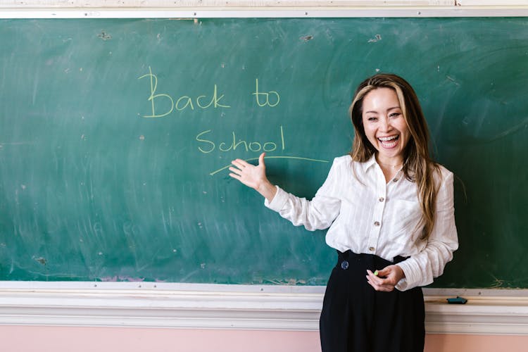 A Happy Teacher Standing Near The Chalk Board While Showing The Message