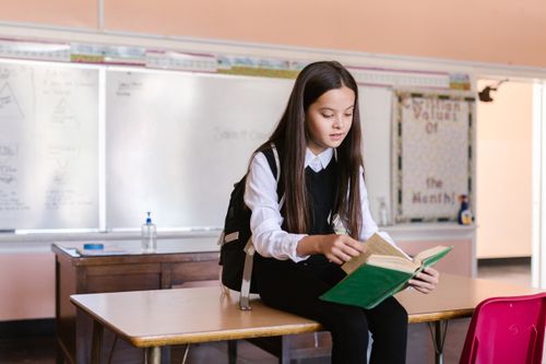 Free Young girl sitting on desk in classroom reading a book, wearing a backpack. Stock Photo